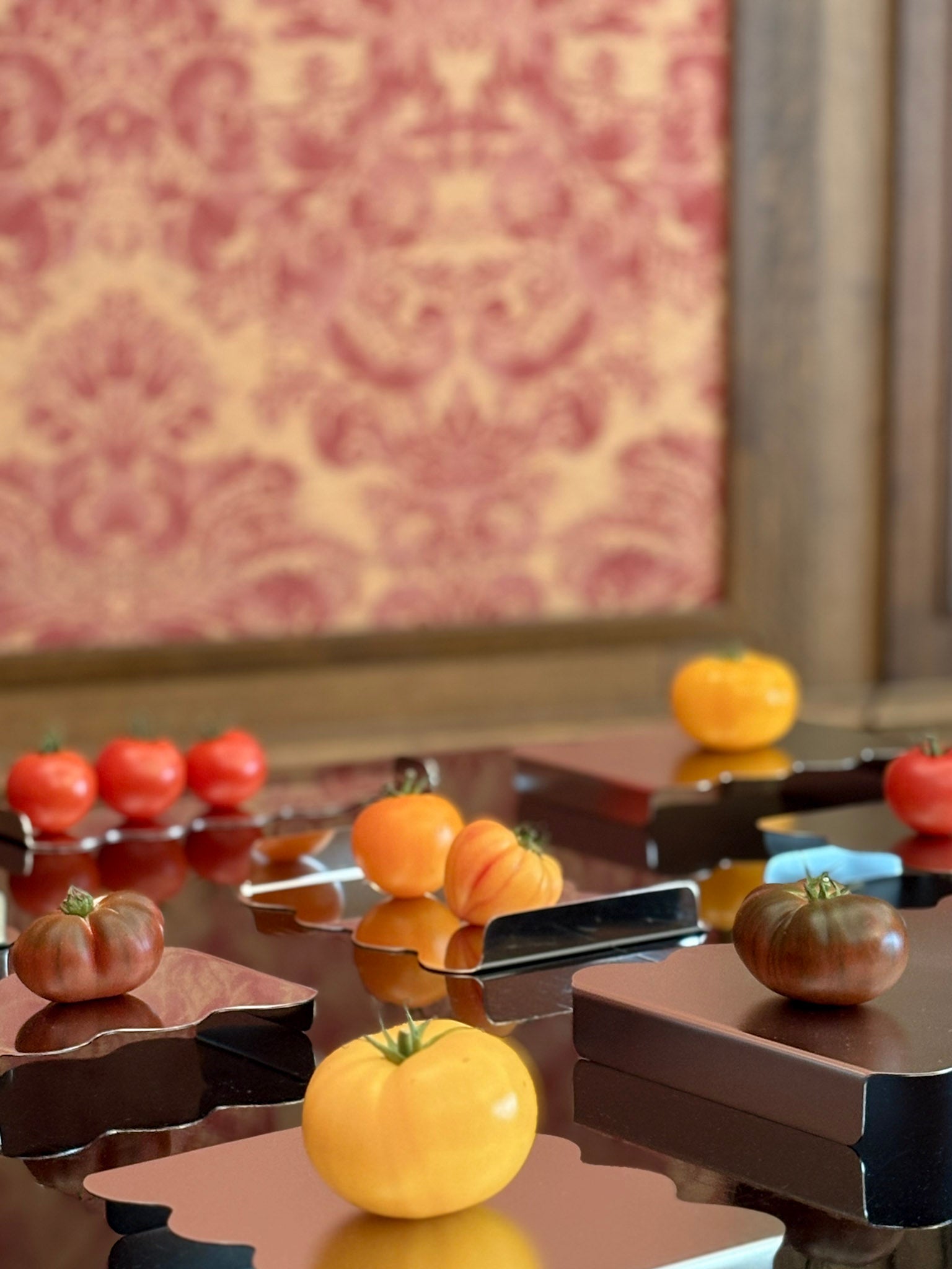 A styled arrangement of stainless steel trays and serving stands with wavy edge with assorted tomatoes, including yellow, red, orange, and dark brown varieties, set against a background of red damask wallpaper and dark wooden furniture.