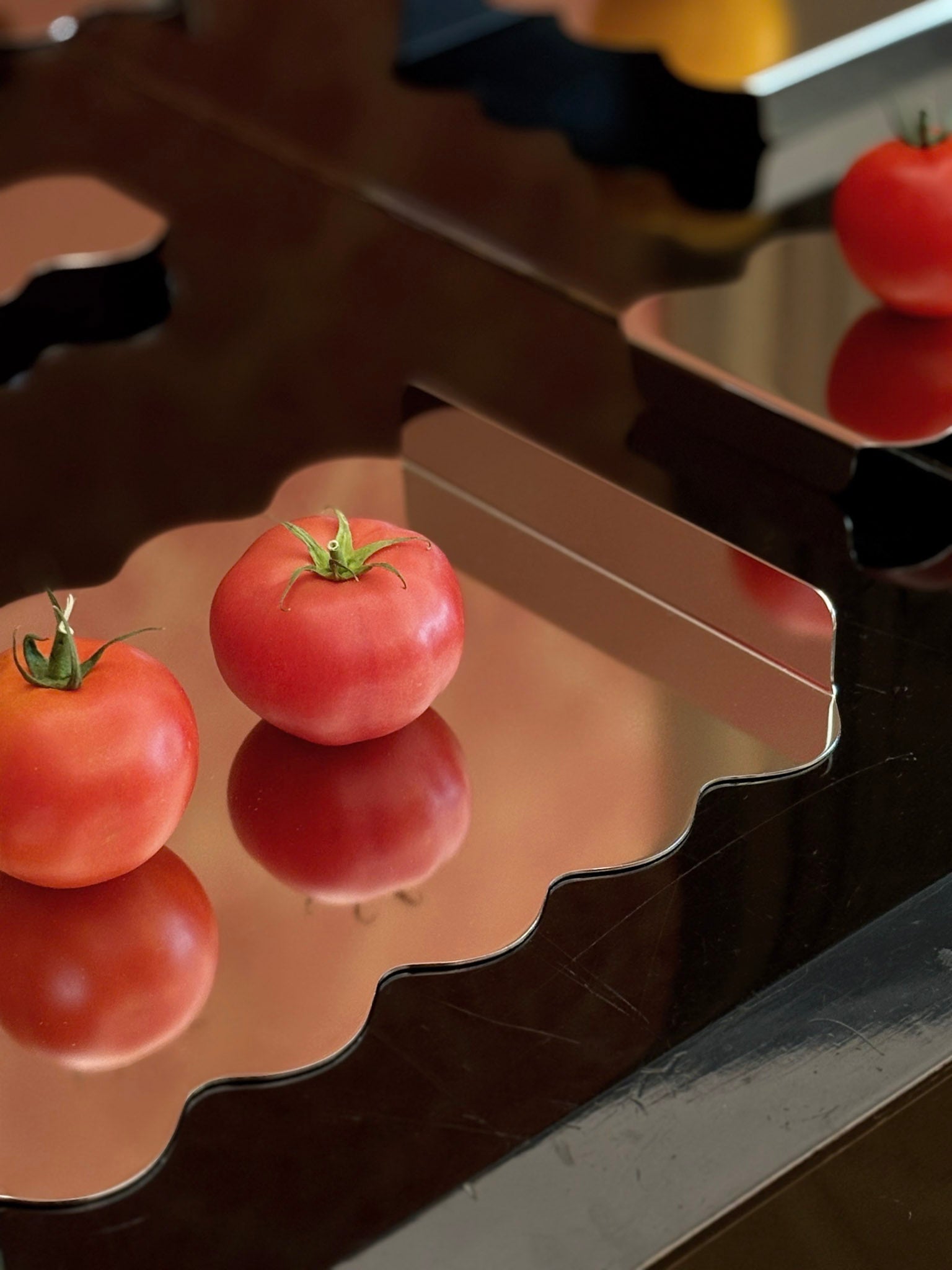 A close-up of a wavy-edged metallic tray holding two red tomatoes, with their reflections visible on the dark reflective surface below.