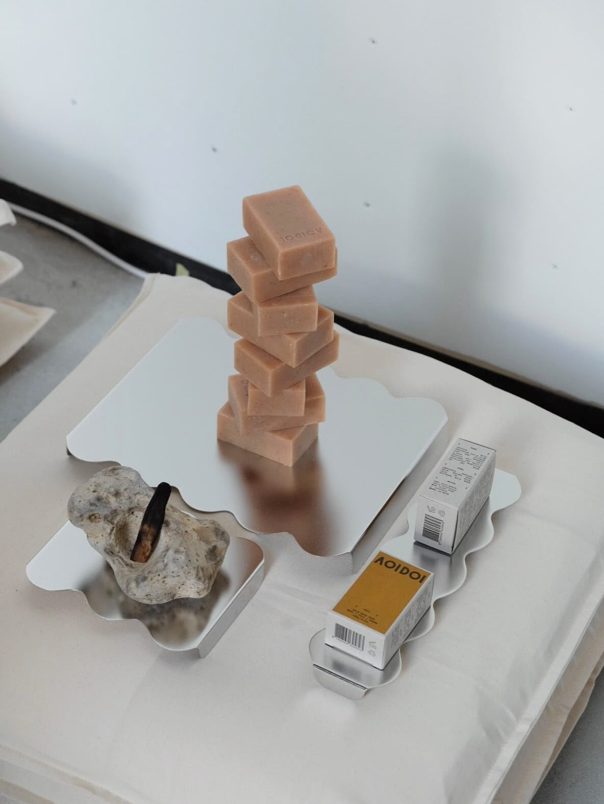 A styled product shot featuring a stack of light peach-pink rectangular bars of soap on a silver scalloped tray, alongside a decorative stone and two Aoidoi soap boxes on a white surface against a light gray background.