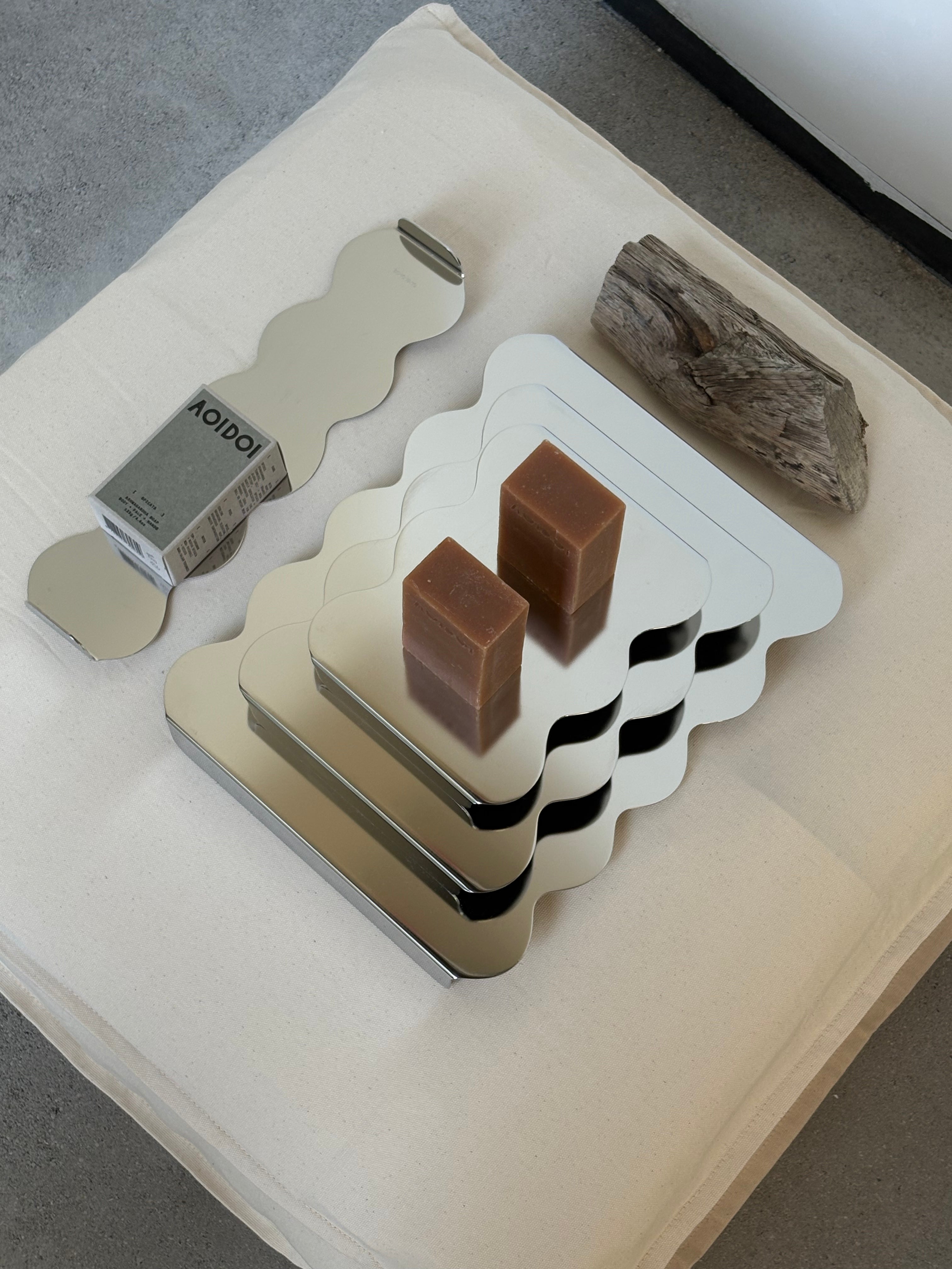 A set of trays with wavy edge and mirror metal finish styled on a cream pillow with two brown soap bars and a piece of driftwood