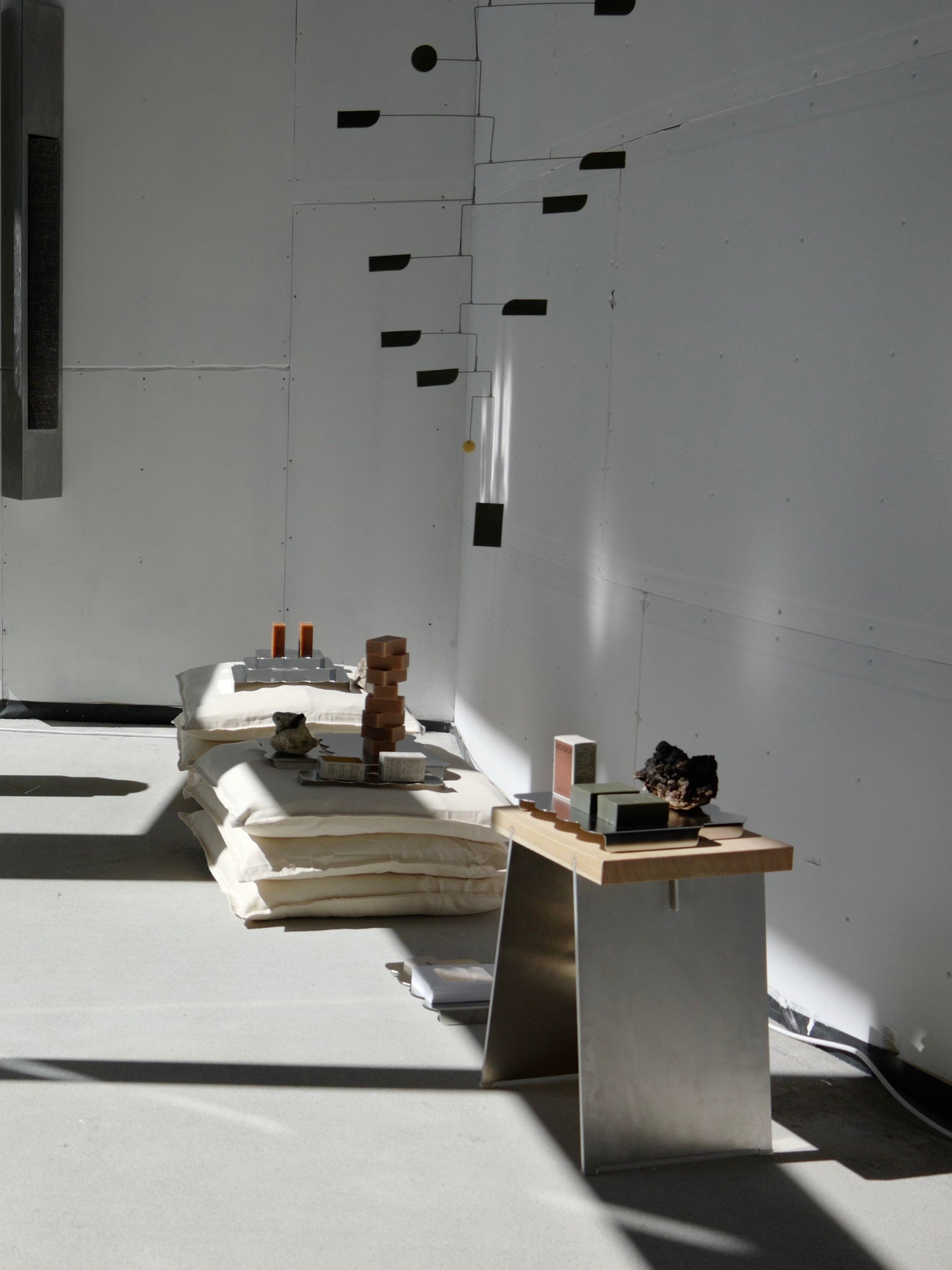 A minimalist interior scene with stacked cream cushions, a small wooden table, and several sculptural objects in a bright white room with strong sunlight and shadows.