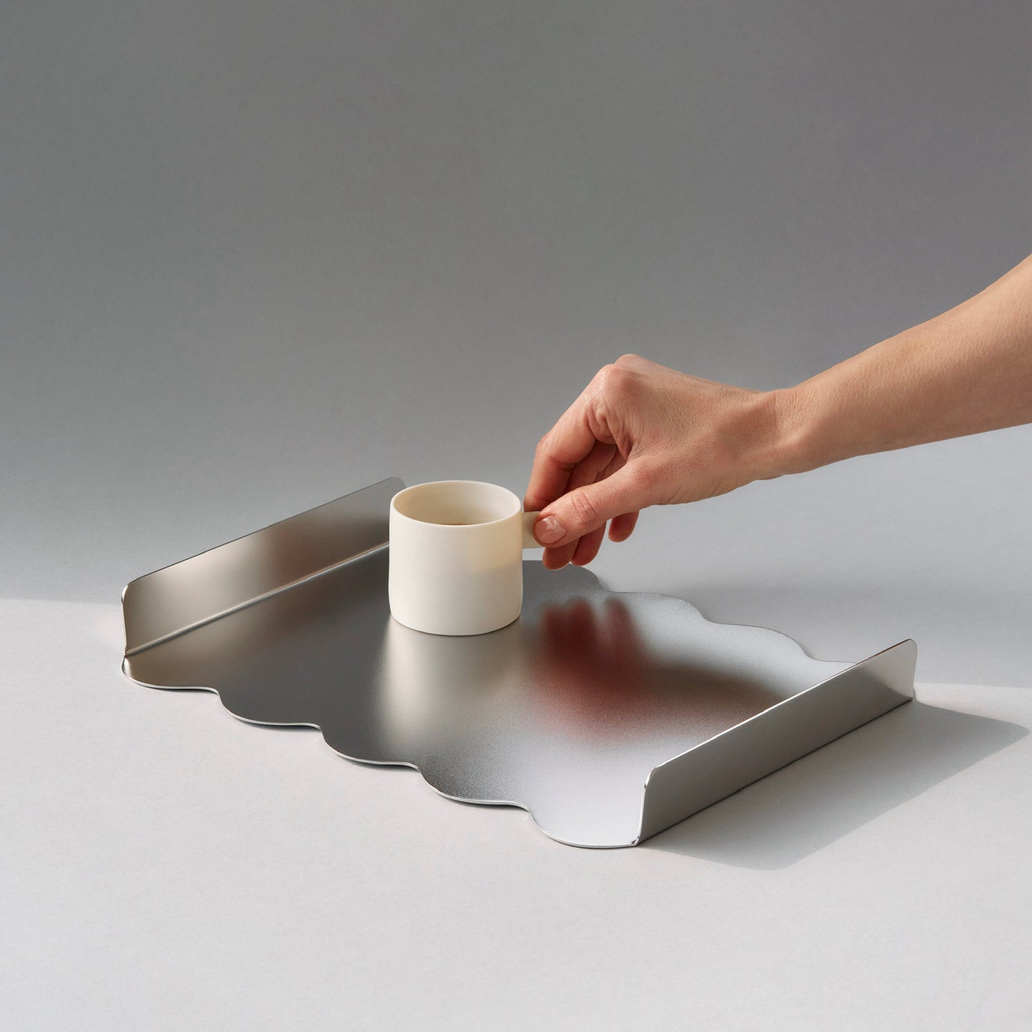 Hand holding a white ceramic cup over a sculptural stainless steel tray with sift matt finish
