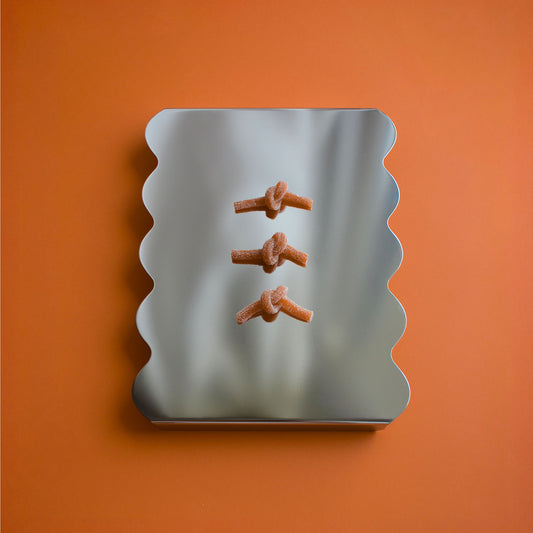 A stainless steel tray with wavy scalloped edges reflecting three small orange knotted candies, set against a solid orange background.