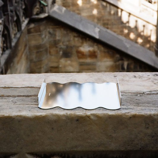 A small metallic silver tray with a wavy, undulating edge sits on a weathered stone ledge, with a blurred stone staircase background.