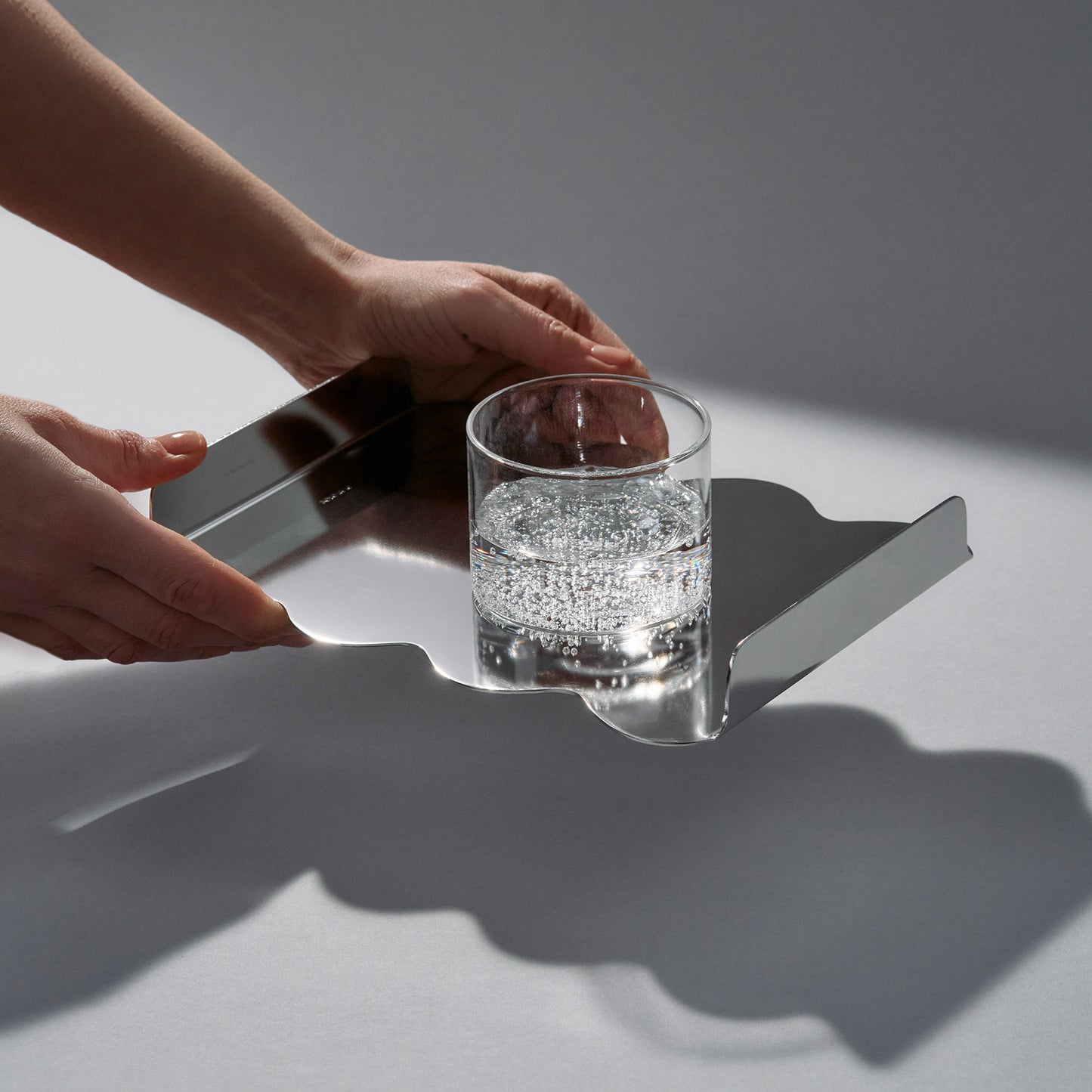 Person holding a mirror sculptural stainless steel tray and a glass of sparklink water