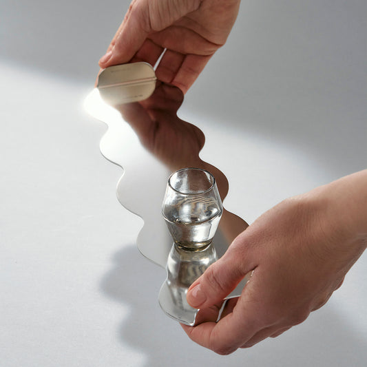 Two hands holding a small glass on a wavy shaped serving tray in reflective stainless steel