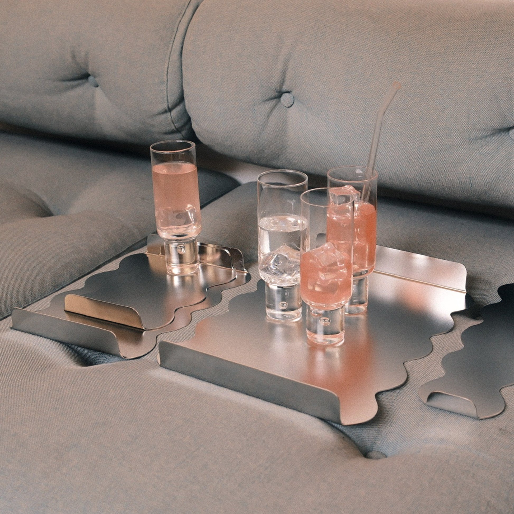 A set of metallic silver trays with wavy cutouts, shown on a gray tufted sofa with several clear shot glasses containing pink drinks and ice.