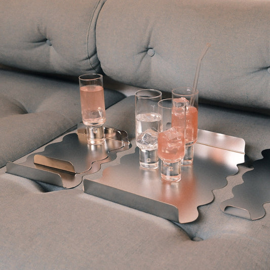 A set of metallic silver trays with wavy cutouts, shown on a gray tufted sofa with several clear shot glasses containing pink drinks and ice.