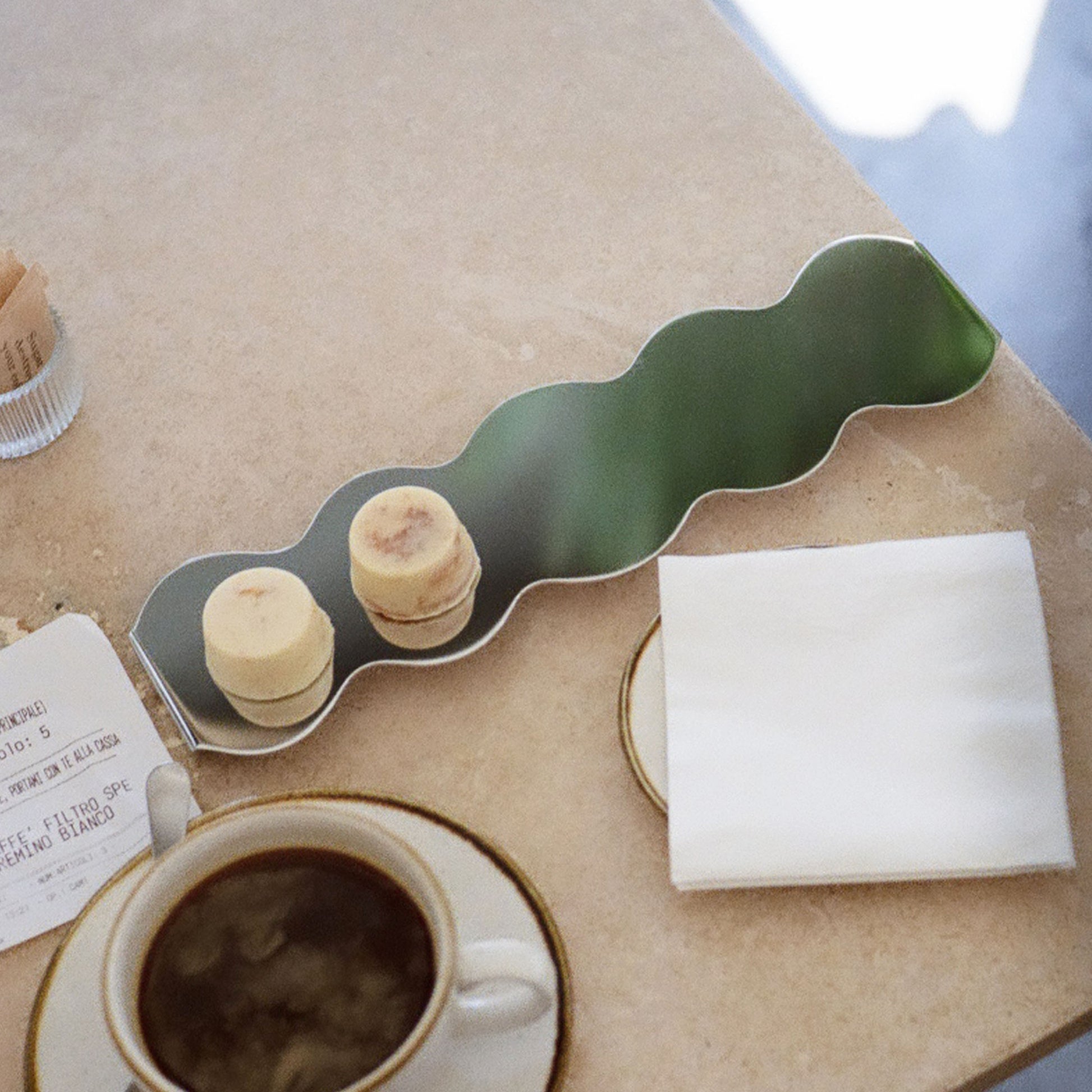 A wavy metal tray serving stand with a green reflective interior, holding two small macarons, styled on a beige tabletop next to a cup of coffee, a napkin, and a receipt.