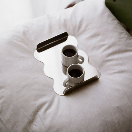 Two small coffee cups on a stainless steel tray with sculptural edges on a white pillow