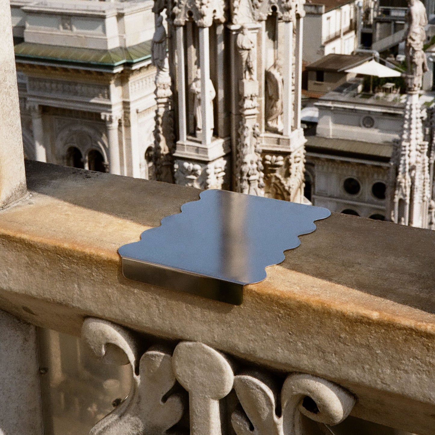 A ectangular metal tray or accessory with a wavy edges resting on a stone balcony railing, with a blurred architectural background of ornate buildings.