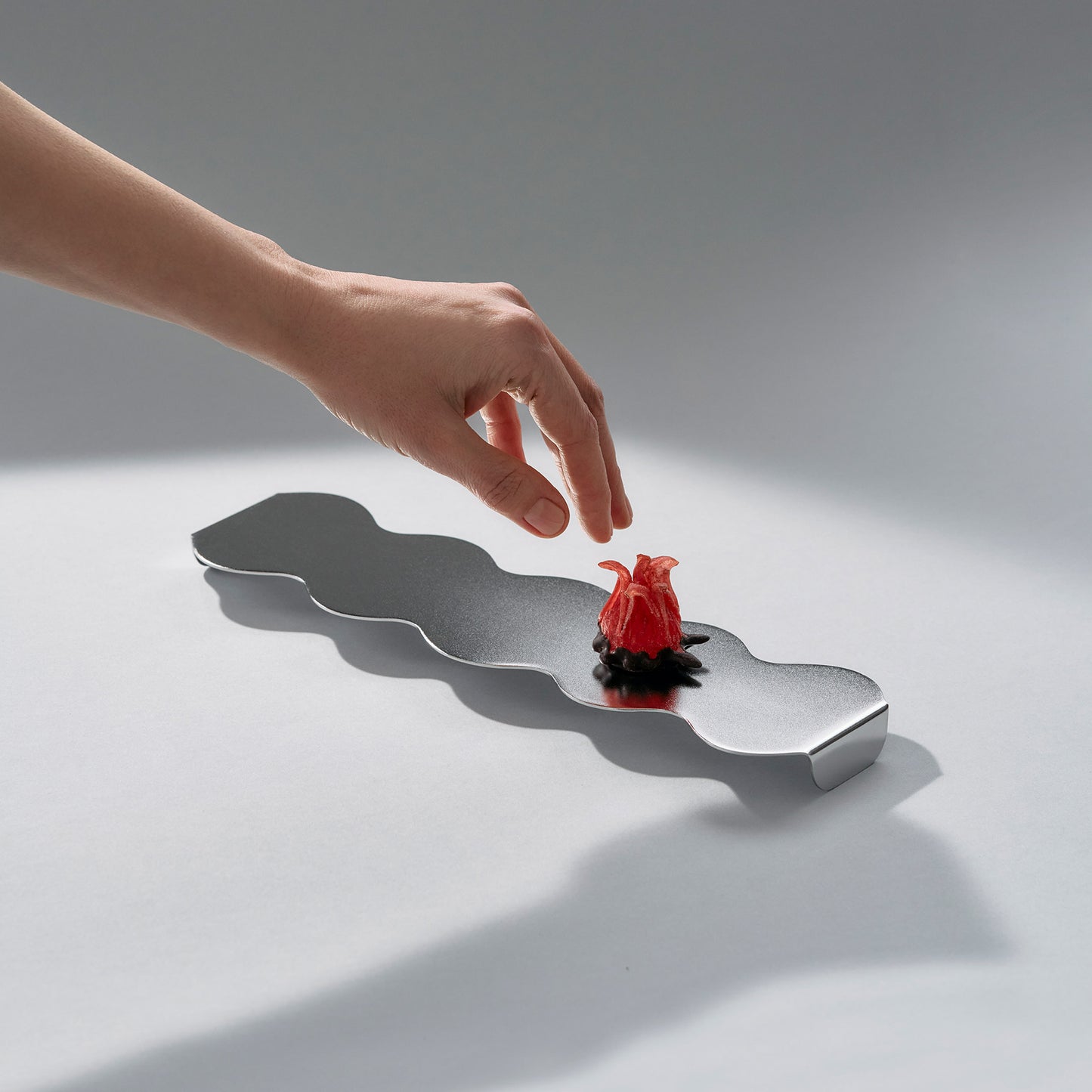 Side view of a sculptural long serving stand in stainless steel and a red candy flower