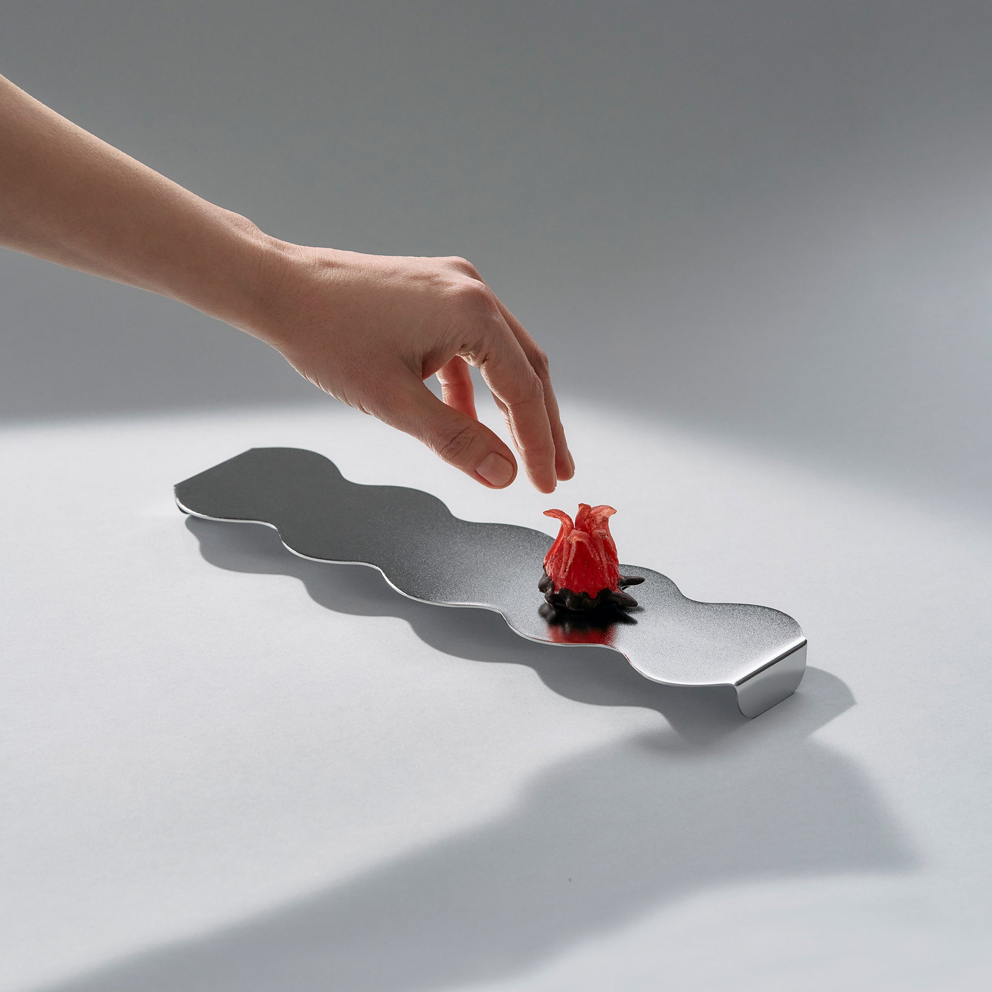 Side view of a sculptural long serving stand in stainless steel and a red candy flower