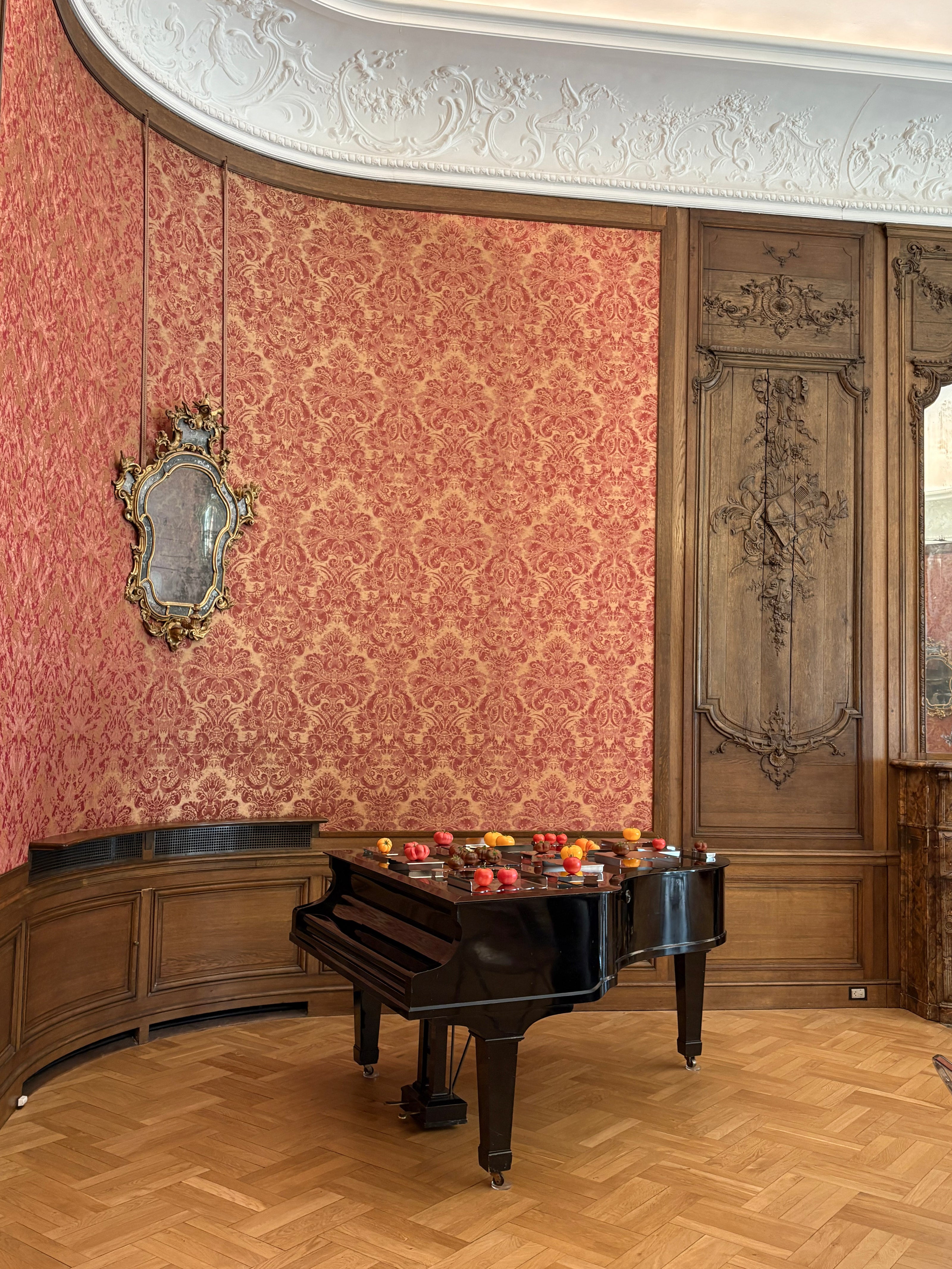 A black grand piano sits in an ornate room with pink damask wallpaper, carved wood paneling, a herringbone wood floor, and a decorative mirror with  metal serving stands and trays with tomatoes.