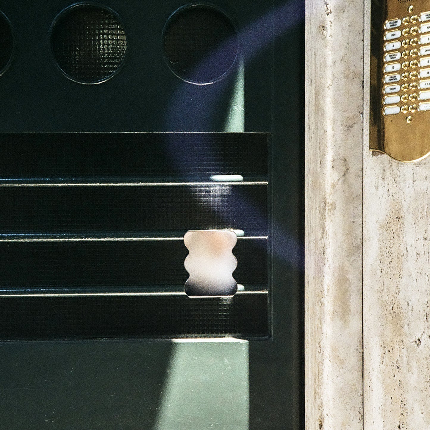 Close-up of a dark textured metal door circular windows, horizontal slats, and a small metal wavy tray centered in the frame, next to a light stone or concrete surface with a gold-toned strip of small rectangular elements.
