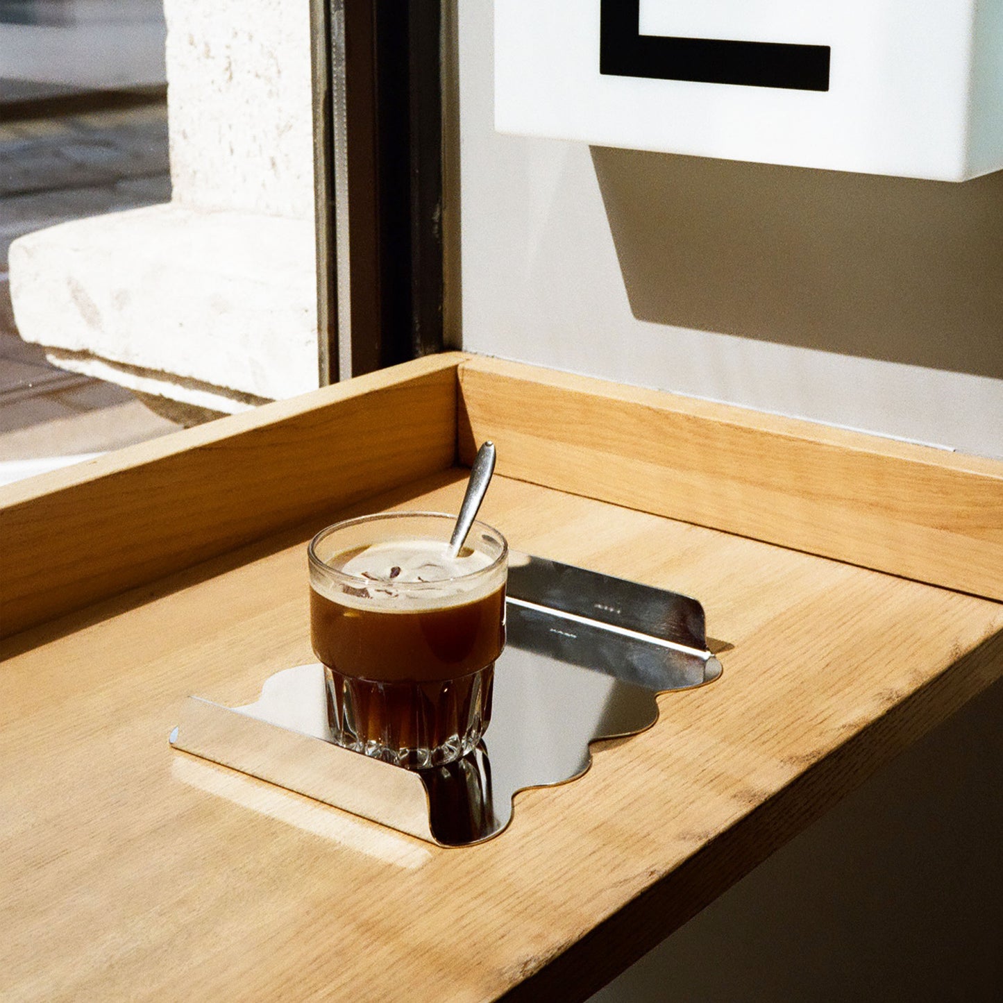 A organic wavy shape metal tray, shown on a light wood table next to a glass of iced coffee with a spoon.